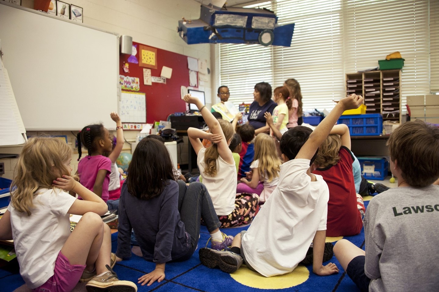 Children sitting on a classroom floor with their arms raised to answer questions as they listen to a teacher.